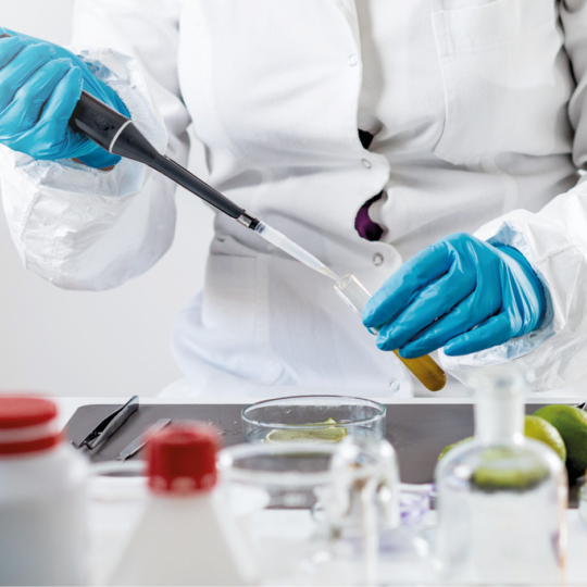 Ein Wissenschaftler im Laborkittel und mit blauen Handschuhen füllt mit einer Pipette Flüssigkeit in ein Reagenzglas, umgeben von verschiedenen Laborgeräten und Behältern auf einem weißen Tisch.