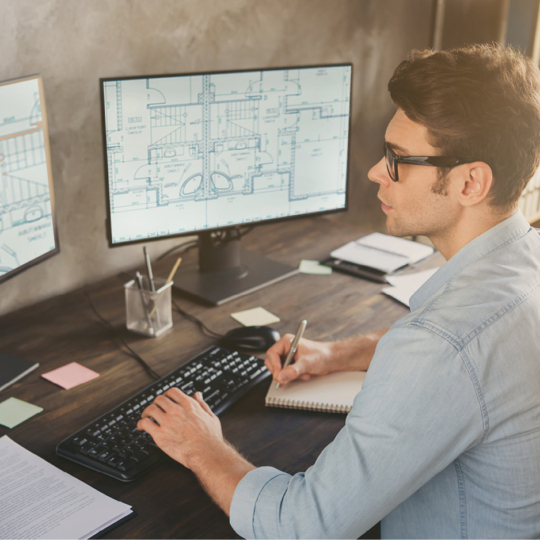 Ein Mann sitzt an einem Schreibtisch und arbeitet an architektonischen Entwürfen, die auf zwei Computermonitoren angezeigt werden, und macht sich Notizen in einem Notizbuch. Der Arbeitsbereich umfasst eine Tastatur, eine Lampe und verstreute Haftnotizen.