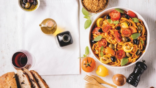 Eine bunte Schüssel mit Nudeln, Tomaten, Oliven und Basilikum auf einem weißen Tisch, umgeben von Brot, Olivenöl, Essig, einem Glas Rotwein, einer Pfeffermühle, Holzbesteck und einer Schüssel mit Bröseln.