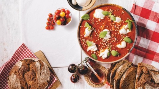 Eine Schüssel Tomatensuppe mit frischem Basilikum, Ricotta und Sonnenblumenkernen steht auf einem Tisch mit geschnittenem Schwarzbrot, Kirschtomaten, Mozzarellakugeln und einer rotkarierten Serviette.
