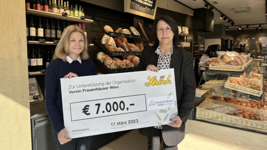 Zwei Frauen stehen in einer Bäckerei und halten einen großen Scheck über 7.000 Euro zur Unterstützung des "Vereins Frauenhäuser Wien" in der Hand. Im Hintergrund sind Brote und Gebäck zu sehen. Der Scheck ist auf den 17. März 2023 datiert.