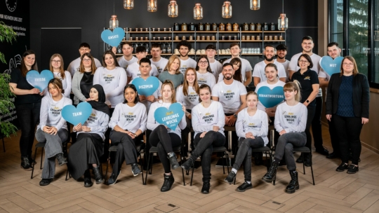 Eine Gruppe von etwa 30 Personen posiert in einem Innenraum, einige sitzend, einige stehend. Mehrere Personen halten blaue herzförmige Schilder mit Aufschriften wie Team und Ehrlich. Im Hintergrund hängen Regale mit Gläsern und Lichtern.