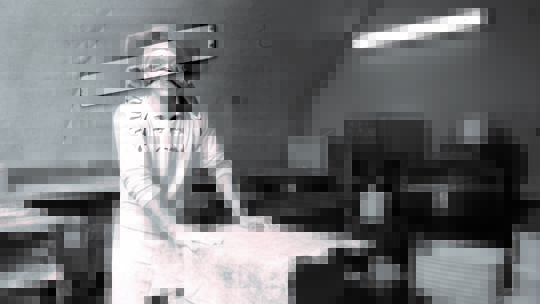 Eine lächelnde Person mit Kochmütze und Schürze steht an einem Tresen in einer Bäckereiküche, umgeben von Backgeräten und gedeckten Tischen.