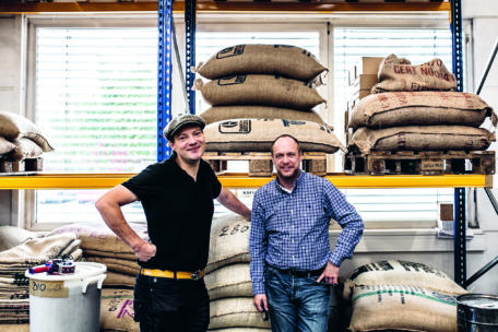 Zwei Männer stehen lächelnd in einem Lagerhaus, hinter ihnen stapeln sich große Jutesäcke auf Paletten. Ein Mann trägt eine Mütze und ein schwarzes Hemd, der andere ein blaukariertes Hemd. Die Umgebung scheint mit Kaffee oder ähnlichen Waren zu tun zu haben.