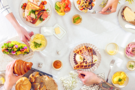 Ein heller Brunch-Tisch mit Waffeln, Toast, Avocado-Sandwich, Croissants, Obst, Rührei, Käse und Kaffee. Mehrere Hände greifen nach dem Essen, das auf einer weißen Tischdecke steht.