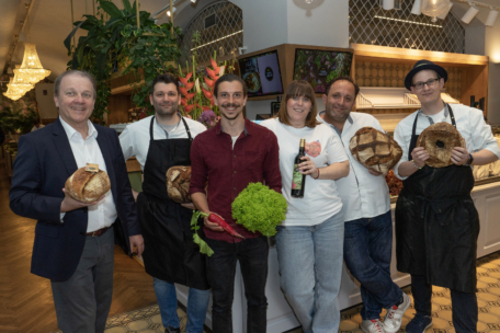 Sechs lächelnde Menschen stehen in einem Raum; drei halten runde Brote, einer hält einen Salat, einer eine Flasche Wein und einer trägt einen Blazer. Die Umgebung scheint eine moderne Bäckerei oder ein Restaurant zu sein.