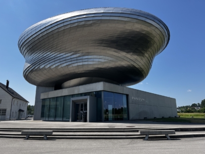 Ein modernes Gebäude mit einer einzigartigen, metallischen, spiralförmigen Struktur an der Spitze und einer Glaswand im unteren Bereich. Auf dem Schild steht Paneum. Im Hintergrund sind blauer Himmel und grünes Gras zu sehen, im Vordergrund Bänke.