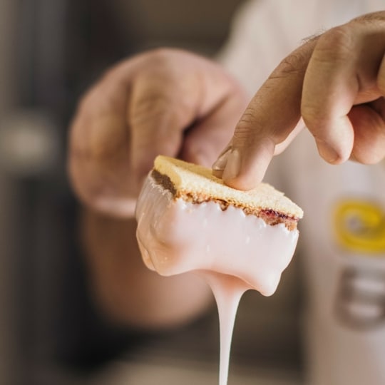 Eine Person taucht einen geschichteten Keks in weiße Glasur und hält ihn vorsichtig mit den Fingern fest, während die Glasur abtropft. Der Fokus liegt auf dem Keks und den Händen, der Hintergrund ist unscharf.