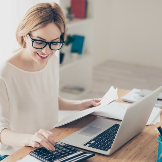 Eine Frau mit Brille sitzt an einem Schreibtisch, lächelt und benutzt einen Taschenrechner. Sie hat Papiere in einer Hand und einen Laptop vor sich. Auf dem Schreibtisch befinden sich außerdem Büromaterial und eine Topfpflanze.