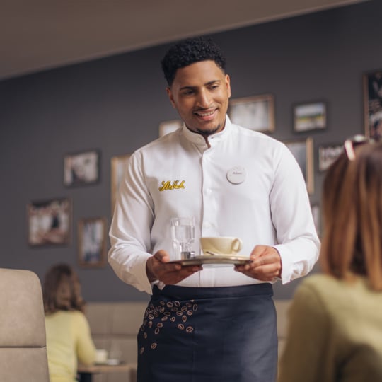 Ein lächelnder Kellner in weißem Hemd und Schürze bedient eine Frau, die an einem Tisch in einem gemütlichen Restaurant sitzt. Er hält ein Tablett mit einer Tasse, einer Untertasse und einem Glas Wasser in der Hand. Gerahmte Fotos schmücken die Wand im Hintergrund.