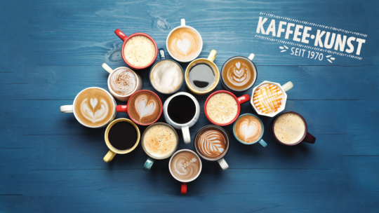 Eine Vielzahl von Kaffeetassen mit verschiedenen Milchkaffees und Kaffeesorten, die in einem Kreis auf einem blauen Holztisch angeordnet sind. Der Text KAFFEE-KUNST SEIT 1970 erscheint in der oberen rechten Ecke.
