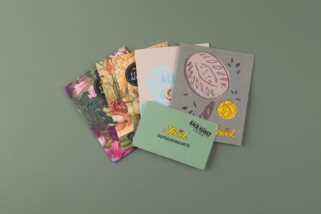 Ein Satz bunter Karten und Umschläge, darunter eine grüne Geschenkkarte mit dem Aufdruck Bäck Gusch Stöck Gutschein, angeordnet auf einer gedeckten grünen Fläche.