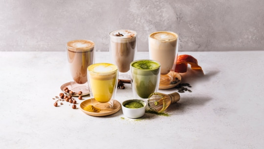 Fünf verschiedene Lattes in klaren Gläsern sind auf einer weißen Fläche arrangiert, darunter die Geschmacksrichtungen Matcha und Kurkuma. Ein Bambusbesen, Matcha-Pulver, Gewürze und kleine Kaffeebohnen lassen eine gemütliche Bäckerei-Atmosphäre aufkommen.