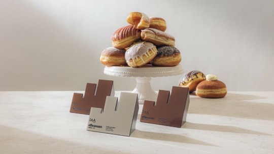 Ein weißer Kuchenständer hält einen Stapel verschiedener Donuts. Davor sind auf einer hellen Fläche drei IAA Effie Awards Trophäen ausgestellt. Ein weiterer Donut steht im Hintergrund.