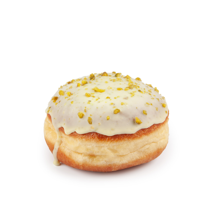 Ein runder, mit weißem Zuckerguss überzogener und mit zerstoßenen Pistazien bestreuter Donut steht auf einem weißen Hintergrund. Etwas Zuckerguss tropft an der Seite herunter.