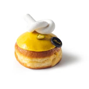 Ein rundes Gebäck mit gelber Glasur, gekrönt von einem weißen Baiser-Twist, einer grünen Pistazie und einer kleinen schwarzen Schokoladenscheibe mit goldener Schrift auf weißem Grund.