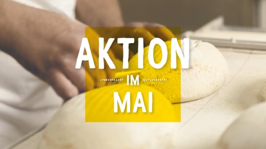 Nahaufnahme eines Bäckers, der Teig formt. Der eingeblendete Text in Deutsch lautet: AKTION IM MAI, wobei die Worte durch einen gelben Hintergrund hervorgehoben werden.