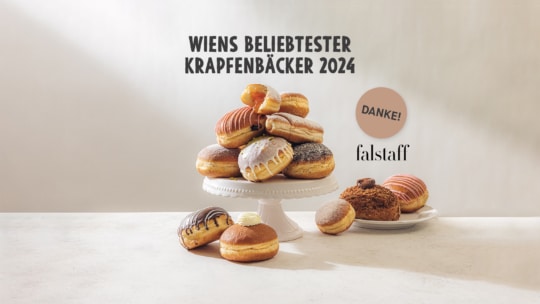 Ein Stapel gefüllter Donuts liegt auf einem weißen Kuchenständer, um den herum weitere Donuts stehen. Darüber steht der Text in Deutsch: Wiens beliebtester Krapfenbäcker 2024. Rechts daneben stehen die Worte Danke! und Falstaff.