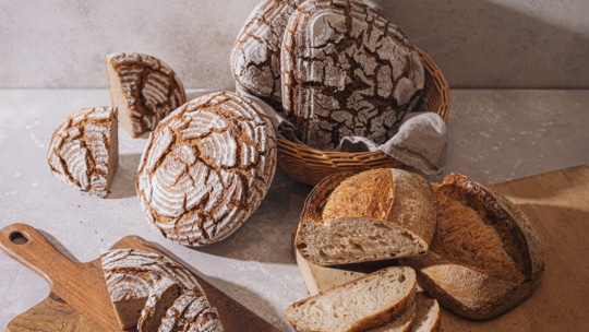 Auf Holzbrettern und in einem Korb sind mehrere runde Laibe mit rustikalem, knusprigem Brot und Brotscheiben ausgestellt, wobei die Krusten leicht mit Mehl bestäubt sind. Die Szene ist vor einem neutralen Hintergrund gehalten.
