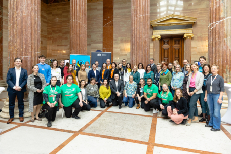 Eine große Gruppe von Menschen posiert in einer großen Halle mit Marmorsäulen und kunstvollem Dekor. Mehrere Personen im Vordergrund tragen leuchtend grüne Hemden, während andere in Geschäfts- oder Freizeitkleidung gekleidet sind.
