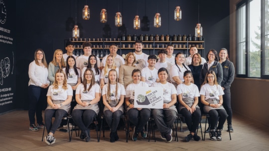 Eine Gruppe von Menschen posiert in einem Innenraum in zwei Reihen, die meisten tragen weiße Hemden mit einem Logo. Einige sitzen, während andere hinter ihnen stehen. Sie lächeln, und im Hintergrund sind Hängelampen und Regale mit Gläsern zu sehen.