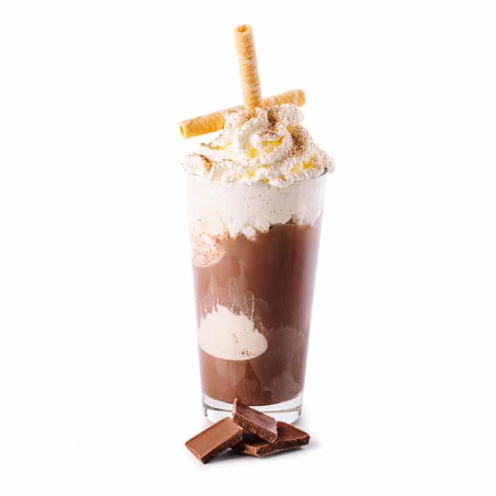 Ein hohes Glas Schokoladenmilchshake mit Schlagsahne, zwei gerollten Waffelstäbchen und Schokoladenraspeln, mit Schokoladenstückchen vor dem Glas auf weißem Hintergrund.