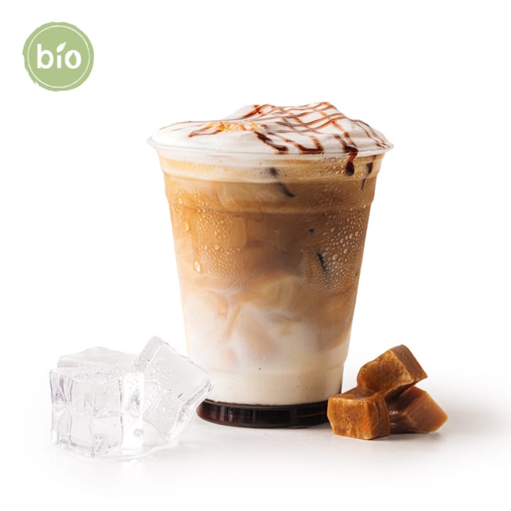 Geeister Karamell-Macchiato in einem durchsichtigen Plastikbecher mit Schaum und Karamellspritzern obenauf, neben zwei Eiswürfeln und Karamellbonbonstücken, auf weißem Hintergrund. Ein grünes Bio-Etikett erscheint in der oberen linken Ecke.
