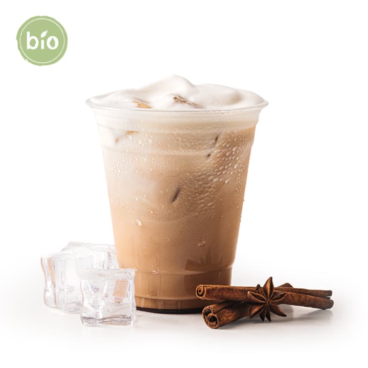 Ein Plastikbecher mit geeistem Milchtee, der mit Schaum bedeckt ist, neben drei Eiswürfeln, zwei Zimtstangen und einem Sternanis. Ein grünes "Bio"-Etikett erscheint in der oberen linken Ecke. Der Hintergrund ist weiß.
