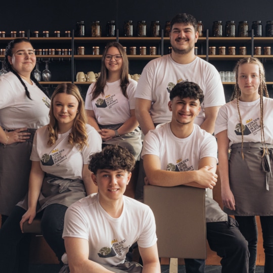 Sieben junge Erwachsene in passenden weißen T-Shirts und grauen Schürzen posieren und lächeln gemeinsam in einem modernen Café, hinter dem Regale mit Gläsern und Gläsern stehen.
