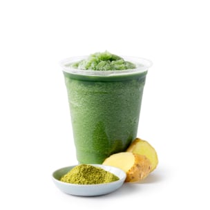 Ein Plastikbecher, gefüllt mit einem grünen Slushy-Getränk, steht neben einer kleinen Schale mit grünem Pulver und zwei Stücken frischem Ingwer auf einem weißen Hintergrund.