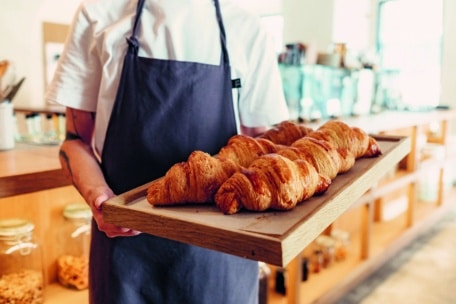 Eine Person mit weißem Hemd und dunkler Schürze hält ein Holztablett mit mehreren frisch gebackenen Croissants in einer hellen, modernen Bäckerei.