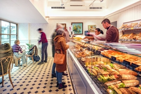 Eine helle Bäckerei mit Menschen, die an einer Glasvitrine mit Gebäck und belegten Brötchen bestellen; andere sitzen an Tischen und unterhalten sich am Fenster.