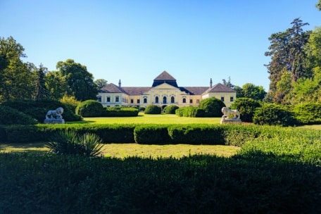 Ein prächtiges gelbes Herrenhaus mit einem zentralen Bogen und symmetrischen Flügeln ist von gepflegten Hecken, üppigen grünen Rasenflächen und zwei steinernen Löwenstatuen umgeben, die sich vor einem klaren blauen Himmel abzeichnen.