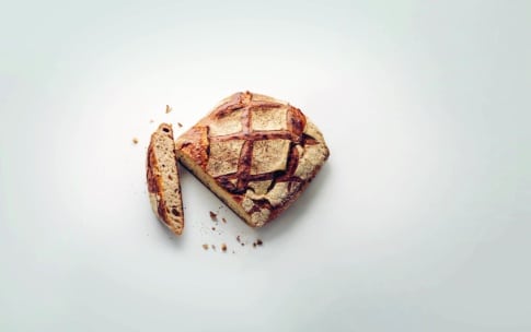 Ein rustikaler, quadratischer Brotlaib mit einem Kreuzmuster auf der Oberseite liegt auf einer weißen Fläche. Eine Scheibe ist abgeschnitten und liegt neben dem Laib, daneben liegen ein paar verstreute Krümel.