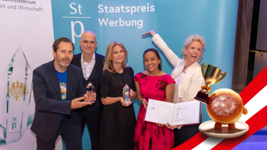 Fünf Personen stehen lächelnd, mit Trophäen und einer Urkunde in der Hand, vor Bannern für den Staatspreis Werbung. Im Vordergrund erscheint ein Cartoon-Gebäck mit einer Trophäe auf einem rot-weiß gestreiften Hintergrund.