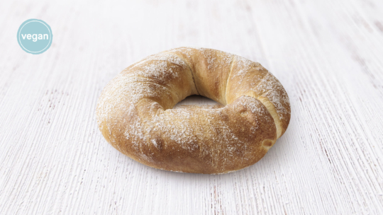 Ein einzelnes, rundes, goldbraunes, mit Puderzucker bestäubtes Croissant liegt auf einer weißen Holzfläche. Auf einem blauen Kreisetikett in der Ecke steht "vegan".