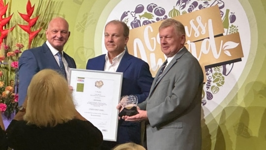 Drei Männer in Anzügen posieren für ein Foto bei einer Preisverleihung und halten eine gerahmte Urkunde und eine durchsichtige Trophäe. Eine Frau steht davor und fotografiert sie. Der Hintergrund ist mit einem Logo und dekorativen Pflanzen geschmückt.