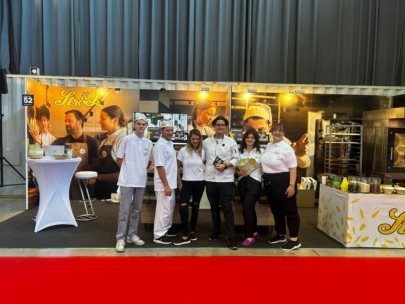 Sechs Personen stehen lächelnd vor einem Bäckereistand mit Werbebannern, einem Tisch und Backgeräten. Sie tragen weiße Uniformen oder Hemden, und der Schauplatz scheint eine Veranstaltung oder Ausstellung zu sein.