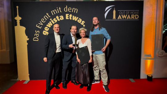 Vier Personen stehen auf einem roten Teppich vor einer schwarz-goldenen Kulisse mit der Aufschrift Das Event mit dem gewissen Etwas. OUT OF HOME AWARD. Drei von ihnen halten gerahmte Auszeichnungen in der Hand und lächeln in die Kamera.