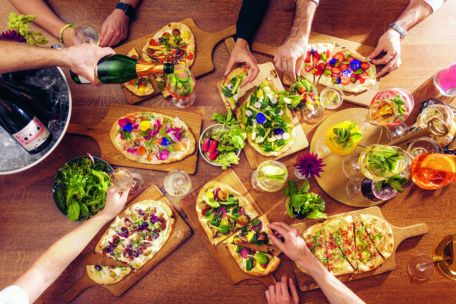 Eine Gruppe von Menschen teilt Fladenbrote, Salate und Getränke an einem Holztisch. Die Hände greifen nach dem Essen und jemand schenkt Sekt ein. Bunte Teller und Gläser schaffen eine lebendige, festliche Atmosphäre.