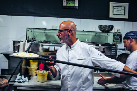 Ein Koch in einem weißen Kittel hält eine Pizza auf einem hölzernen Schieber in einer belebten Küche und bereitet sich darauf vor, sie in einen Ofen zu schieben. Eine andere Person arbeitet neben ihm, im Hintergrund sind Zutaten und Küchengeräte zu sehen.