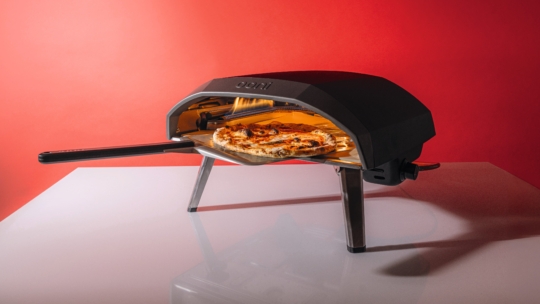 Ein schwarzer Pizzaofen mit dem Markennamen Ooni auf der Oberseite backt eine Pizza auf einer Schaufel, wobei die Flammen im Inneren sichtbar sind. Der Ofen steht auf einer weißen Fläche vor einem roten Hintergrund.