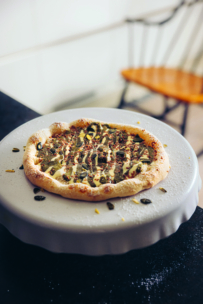 Eine frisch gebackene, mit Kernen und Soße belegte Pizza liegt auf einem weißen Kuchenständer, während im unscharfen Hintergrund ein Holzstuhl und ein Teil eines Tisches zu sehen sind.