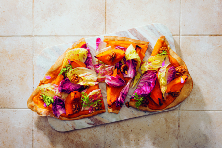 Geschnittenes Fladenbrot, belegt mit buntem Salat, gerösteter orangefarbener Süßkartoffel, Walnüssen und Kräutern, serviert auf einem Marmorbrett auf einer gefliesten Oberfläche.