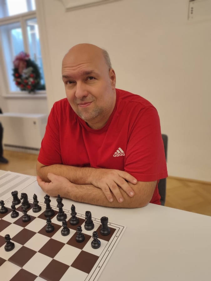Ein Mann in einem roten Adidas-Hemd sitzt an einem Tisch mit einem Schachbrett vor ihm, die Arme verschränkt und in die Kamera blickend. Die Umgebung scheint ein Innenraum mit einem Fenster und einem Blumenkranz im Hintergrund zu sein.