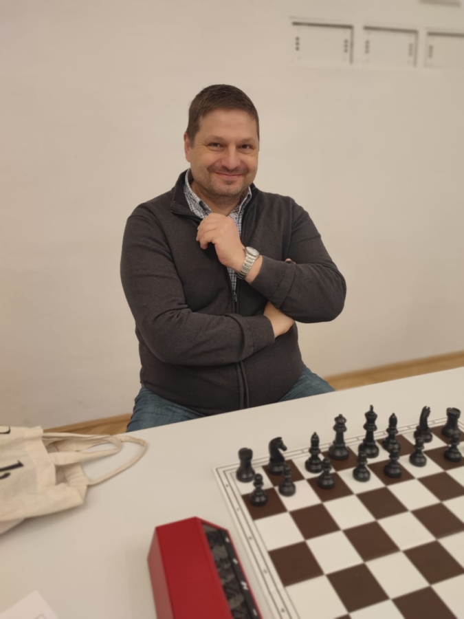 Ein Mann sitzt an einem Tisch mit einem Schachbrett vor sich und lächelt in die Kamera. Er trägt eine dunkle Jacke mit Reißverschluss und blaue Jeans. Eine beigefarbene Tasche und eine rote Schachuhr liegen ebenfalls auf dem Tisch.