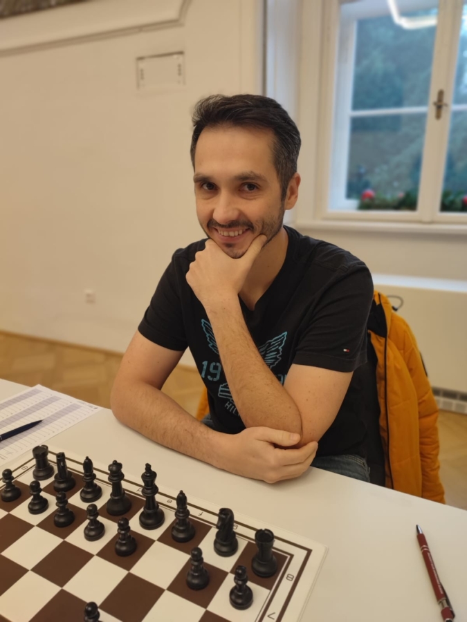 Ein Mann mit dunklem Haar und Bart lächelt, während er an einem Tisch mit einem Schachbrett sitzt und sein Kinn auf seine Hand stützt. Er trägt ein schwarzes T-Shirt und eine gelbe Jacke, die über den Stuhl drapiert ist. Im Hintergrund ist ein Fenster zu sehen.