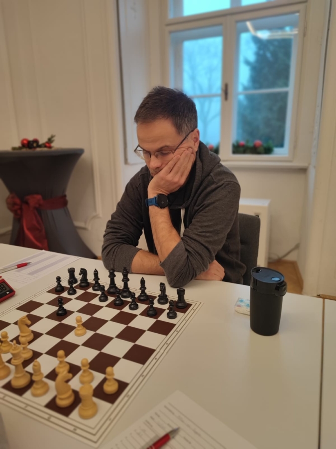 Ein Mann, der eine Brille und einen grauen Pullover trägt, spielt nachdenklich Schach an einem Tisch in einem Haus. Die Schachfiguren sind auf dem Brett angeordnet; auf dem Tisch liegen Spielzettel, eine Tasse und ein Stift. Im Hintergrund sind ein Fenster und ein dekorierter Tisch zu sehen.
