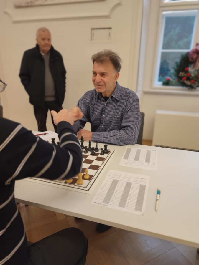 Zwei Männer spielen Schach an einem Tisch in einem Haus, mit Spielberichtsbögen und einem Stift in der Nähe. Ein Mann lächelt, während eine andere Person im Hintergrund in der Nähe eines Fensters mit einem Blumengesteck steht.