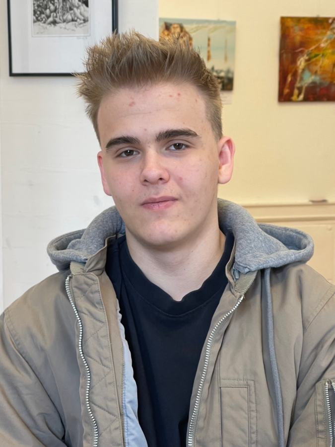 Eine junge Person mit kurzen, hellbraunen Haaren, die eine hellbraune Jacke über einem grauen Kapuzenpulli und einem schwarzen Hemd trägt, sitzt in einem Raum, in dem Kunstwerke und Fotos an einer weißen Wand im Hintergrund zu sehen sind.